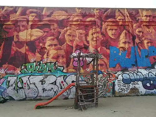 A brick wall depicting a mass of people with raised fists. It has been defaced wit graffiti. A slide for children is in front of it.