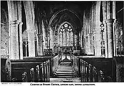 The interior of a building, symmetric with two lines of tall pillars on either side, with candelabras and wooden benches between them.