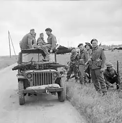 A black-and-white photograph of a jeep with soldiers sitting on top and standing beside it. The soldiers sitting on the jeep are three German soldiers and one British soldier who is interrogating the Germans. On the bonnet of the jeep is small motorcycle, while in the background is a Horsa glider