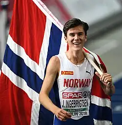 Photo of Jakob Ingebrigtsen in a track outfit while carrying a short flagpole with the Norwegian flag over his shoulder