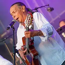 Canut Reyes wearing a white shirt, playing an acoustic guitar and singing into a microphone onstage