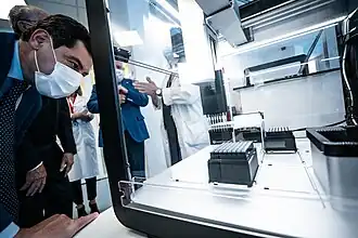 male presenting individual with a white mask over their mouth and nose looking at the insides of the liquid handling robot