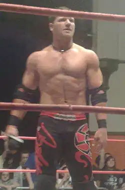 A white male wearing black and red wrestling gear standing in a wrestling ring.