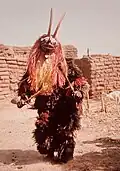 A dancing man-woman in an orange antelope mask with two horns.