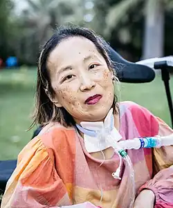 Alice Wong, a woman wearing a colorblock pink and orange top, sits in her wheelchair in a park on a sunny day