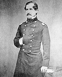 A three-quarters length monochrome photograph of a white man in a dark military uniform; he has dark hair and mustache, his right hand is tucked into the breast of his uniform, and he is looking to the camera's left.
