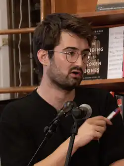 Headshot of Adam Aleksic speaking at a book event. He is a young, white man with dark, brown hair and a full beard, wearing round glasses and a simple, black T-shirt.