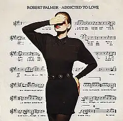A model with a hand raised covering her eyes, her other hand is on her hip. She is wearing a black dress and dark tights. Her face is heavy with makeup, the white of her face contrasting dramatically with her red lips. The background is a filled by a sheet of music notation.