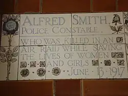 A tablet formed of five tiles of varying sizes, elaborately decorated with flowers and a stylised English policeman's helmet and bordered by yellow and blue flowers in an art nouveau style. The tablet reads "Alfred Smith, Police Constable, who was killed in an air raid while saving the lives of women and girls June 13, 1917".