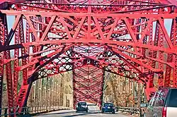A red steel bridge arch is seen from a car going under the metalwork along the bridge deck