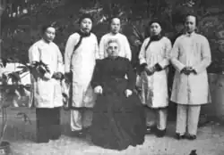 A group portrait of six women, taken outdoors; five Chinese women stand in a row, wearing light-colored tunics and trousers; one white woman, older, is seated in front of the group, wearing all black, with her grey hair dressed in an updo.