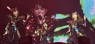 Three women wearing similar costumes consisting of a metallic top and dark skirt, performing on stage