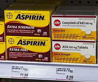 Four boxes of medication on a store shelf above price tags. The two on the left are yellow with "Aspirin" in bold black type and explanatory text in English on the top box and French on the bottom. The two on the right are slightly smaller and white with the word "Life" in the corner inside a red circle. The text, in French on top and English below, describes the medication as "acetylsalicylic acid tablets"