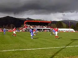 A football match at a football ground. A main stand is visible with hills in the background