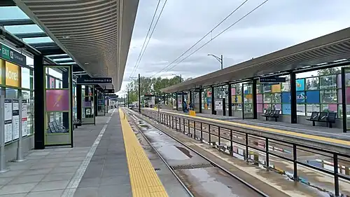 BelRed station on opening weekend in April 2024. The station has two platforms on the sides of the station with two tracks running down the center. There are shelters with glass walls and roofs along the platform, providing protection from the weather. The platform is made of concrete and has yellow lines and railings. There are benches for passengers to wait, and there are signs with information about the station and train schedules. The sky is cloudy, and there are puddles on the track bed.