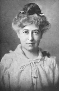 A middle-aged white woman with dark eyes and grey hair; her hair is in a bun on top of her head; she was a nothing buttoned blouse with a tie closure at the neck.