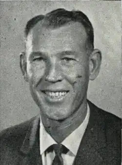 A black and white photograph of a white man, smiling and wearing a suit and tie, shot from the chest up