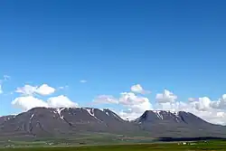mountains with a grassy plain in front of them. A clear blue sky with low, scattered clouds