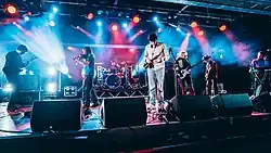 Black Country, New Road performing live on stage in January 2020 under colourful lighting, with all seven band members playing instruments like guitars, drums, a violin, a saxophone and a keyboard.