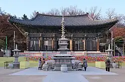 A head-on shot of the Bongeunsa buddhist temple. The temple has a traditional Korean roof and is adorned with various decorative designs and hanja characters. In front of the temple are a few people surrounding a large stone pillar.