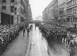 Soldiers with rifles and helmets march along a road past a reviewing party and guard of honour