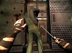 A pair of human arms with brown tape on them holding a screwdriver in the right hand. A man working on a machine, wearing a green jumpsuit and a black shirt, is in front of the arms with blood splatter on the wall to his left.