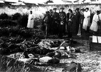 Photograph of several dead bodies lying on the floor. Above them stand members of the police and reporters.