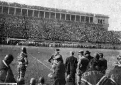 Football players with their backs to the camera; stadium grandstands are visible in the background