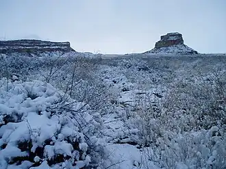 Rocky desert landscape blanketed in snow, shown in near-twilight. Two massifs, several miles in the distance, are snow-covered.