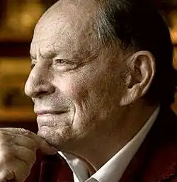 Colour publicity shot, showing profile of an elderly, clean-shaven, smiling man with receding hair, his chin resting on his hand.