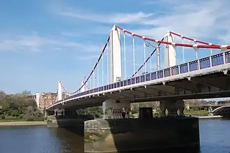 Suspension bridge crossing a wide river, under blue skies with clouds. The main cables are red and the towers white.