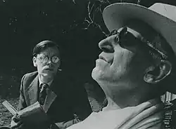 Young white man in spectacles reading to an older (blind) man leaning back in a garden chair.