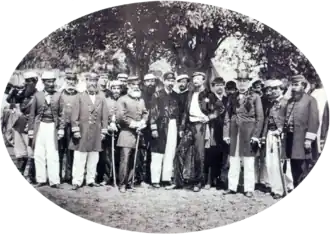 A photograph showing a large group men, most of whom wear uniforms, standing beneath shade trees