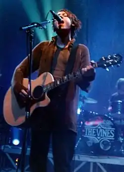 Nicholls is shown singing into a microphone while strumming a guitar.