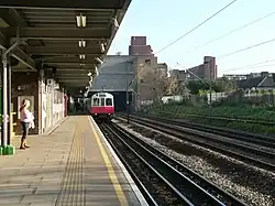 A girl with a pink shirt and blue jeans on a railway platform with a railway track on the right and a white train on it with a red front