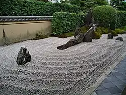 Rock garden with raked gravel and large standing stones in front of a small wall and green plants.