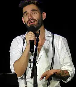 A brown-haired person with a beard and multiple tattoos performing on stage in a white shirt holding a black microphone.