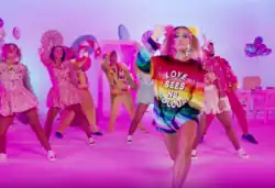 Shot of Delia wearing a multicolored sequined anorak while background dancers are shown in a background decorated with candy.