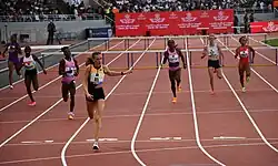Photo of seven athletes seen from the front while running on a red athletics track with hurdles