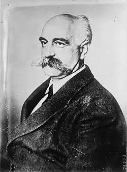 Bald white man, middle-aged, moustached seated, seen in left semi-profile