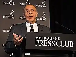 Dellal stands at a lectern giving a presentation. His mouth is open as he speaks and his right hand is held up. He wears a black suit jacket, white shirt and a mostly black tie. Lectern signage and stage backdrop are black with versions of the Melbourne Press Club logo in white.