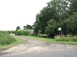 Small farm track leading off a mintor road. behyind a low hedge some single story brick farm buildings peep. A huge tree ominates the right side of the picture