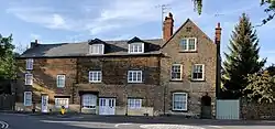 A row of English stone houses with white windows and doors beside a road. There is a stone wall and wooden gate on one side, and several different types of trees around them.