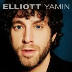 A close-up photo of Elliott Yamin's face against a dark blue background. The artist's name appears above him, with a line in between the first and last name, both colored white.