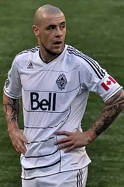 A man wearing a white T-shirt and white shorts, standing on a soccer field with his hands at on his hips, looking to his left.