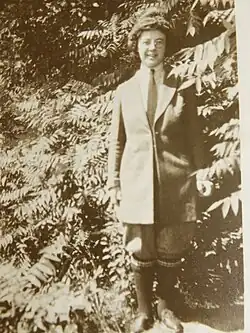 Picture of a woman standing outdoors, wearing jacket, pants, long socks and shoes, with trees and bushes close behind her