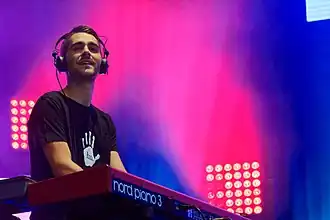 Kevin Rodrigues (Worakls) wearing a black print t-shirt, with a set of large headphones, playing a keyboard onstage and looking out past the camera as if toward an audience