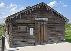 A dark brown log cabin, topped with a pointed roof, seen from the front. Over its main door is a sign saying "Fireweed Studios" in script on white with floral decoration. To its left is a bronze metallic plaque with text explaining the building's history.