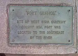 A bronze metallic plaque engraved with the historical description words "Fort Orange: Site of West India Company Colony 1624, was located to the southeast by the river."