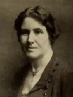 A white woman with short dark hair, wearing a dark collared jacket and a delicate necklace at her throat